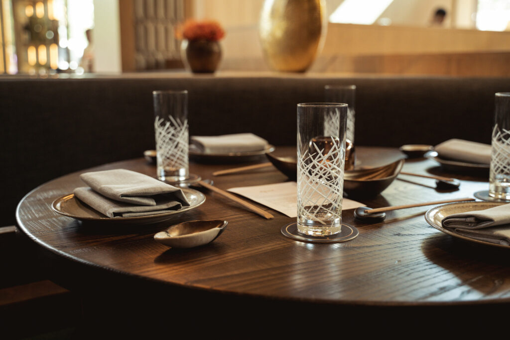 A round dining table set with glasses, napkins, bowls, and a menu in an elegant, softly lit restaurant setting.