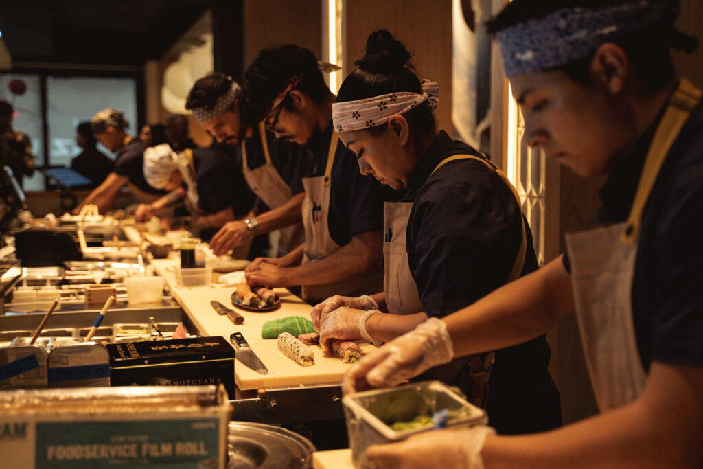 Several chefs in aprons and bandanas prepare food at a sushi counter, focusing intently on their tasks while using various ingredients and utensils.