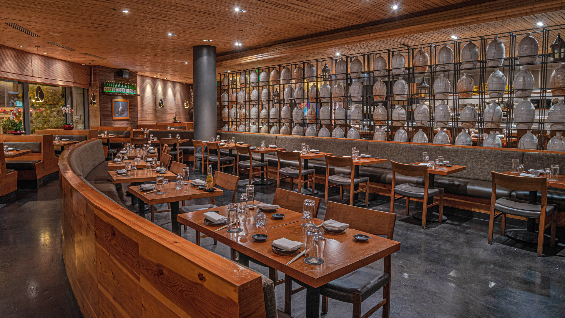 A modern restaurant interior with wooden tables and chairs, set with glasses and plates. A wall of decorative jars and soft lighting create a cozy atmosphere. No people are present.