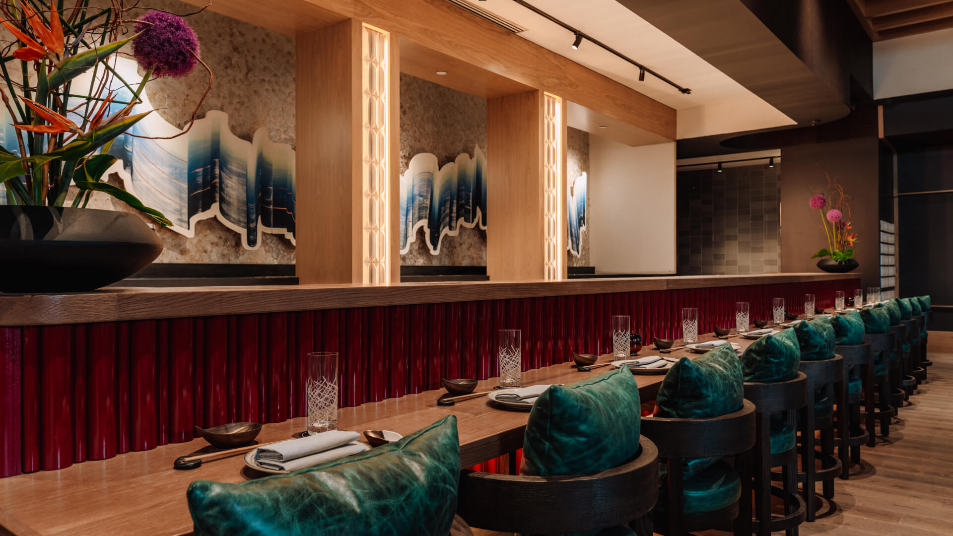 A modern restaurant interior featuring a wooden dining counter with teal-cushioned chairs, decorative plants, and abstract wall art, with soft lighting creating a warm ambiance.